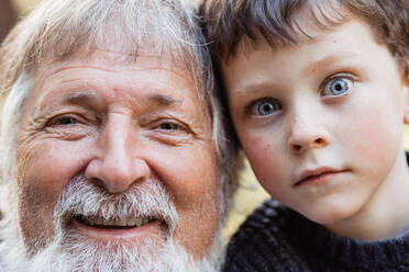 Positiv gealterter Mann mit grauem Bart und Haar und niedliches lächelndes Kind mit blauen Augen, das die Kamera im Wald vor einem unscharfen Hintergrund ansieht - ADSF34230