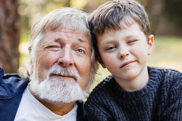Positive aged man with gray beard and hair and cute smiling kid with blue eyes looking at camera in forest against blurred background - ADSF34231