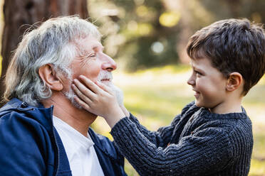Seitenansicht eines fröhlichen kleinen Enkels in einem warmen Pullover, der den grauen Bart eines lächelnden älteren Großvaters mit geschlossenen Augen auf einem unscharfen Hintergrund berührt - ADSF34232