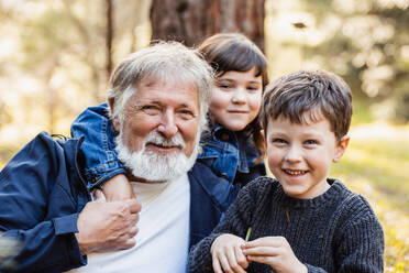 Bärtiger Großvater mit charmanter Enkelin und lachendem Enkel, die im Wald sitzen und in die Kamera schauen - ADSF34233
