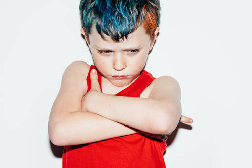 Missgünstiger rebellischer Junge mit gefärbtem Haar, der mit ernstem Gesicht und verschränkten Armen nach unten schaut, während er auf weißem Hintergrund in einem hellen Raum steht - ADSF34237