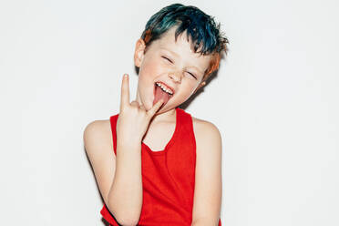 Mischievous boy with colorful dyed hair and closed eyes and showing rock and roll gesture on white background in light room - ADSF34238