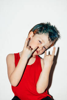 Schelmischer Junge mit bunt gefärbten Haaren zeigt Rock'n'Roll-Geste auf weißem Hintergrund in hellem Raum - ADSF34240