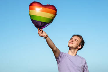 Positiver Transgender-Mann betrachtet herzförmigen Ballon mit LGBT-Symbol in der Hand, während er auf blauem Himmel in der Natur steht - ADSF34242