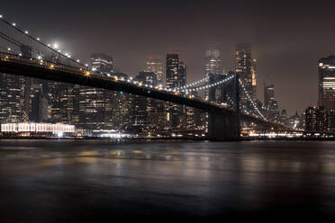 Beleuchtete Brücke über einen ruhigen Fluss gegen moderne Hochhäuser mit leuchtenden Lichtern in New York City bei Nacht - ADSF34258