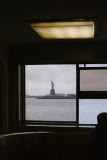 Through window of ferry view of famous Statue of Liberty near sea located in New York city against cloudy sky - ADSF34261