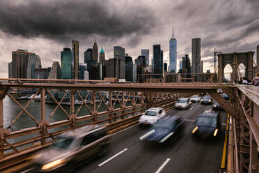 Long exposure of vehicles driving on road on suspension Brooklyn Bridge connecting East River shores in New York City against cloudy gray sky - ADSF34264