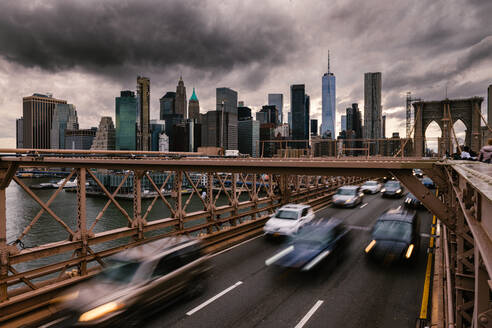 Langzeitbelichtung von Fahrzeugen, die auf der Straße auf der Brooklyn Bridge fahren, die die Ufer des East River in New York City mit dem bewölkten grauen Himmel verbindet - ADSF34264