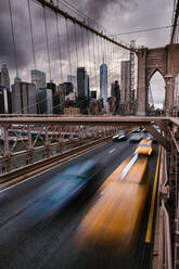 Long exposure of vehicles driving on road on suspension Brooklyn Bridge connecting East River shores in New York City against cloudy gray sky - ADSF34265