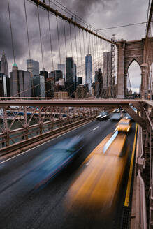 Langzeitbelichtung von Fahrzeugen, die auf der Straße auf der Brooklyn Bridge fahren, die die Ufer des East River in New York City mit dem bewölkten grauen Himmel verbindet - ADSF34265