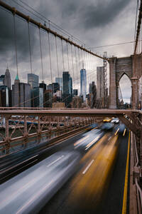 Langzeitbelichtung von Fahrzeugen, die auf der Straße auf der Brooklyn Bridge fahren, die die Ufer des East River in New York City mit dem bewölkten grauen Himmel verbindet - ADSF34266