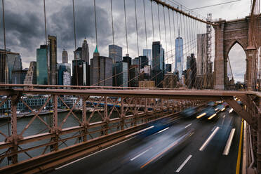 Long exposure of vehicles driving on road on suspension Brooklyn Bridge connecting East River shores in New York City against cloudy gray sky - ADSF34267