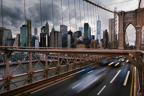 Langzeitbelichtung von Fahrzeugen, die auf der Straße auf der Brooklyn Bridge fahren, die die Ufer des East River in New York City mit dem bewölkten grauen Himmel verbindet - ADSF34267