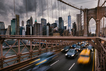 Long exposure of vehicles driving on road on suspension Brooklyn Bridge connecting East River shores in New York City against cloudy gray sky - ADSF34268
