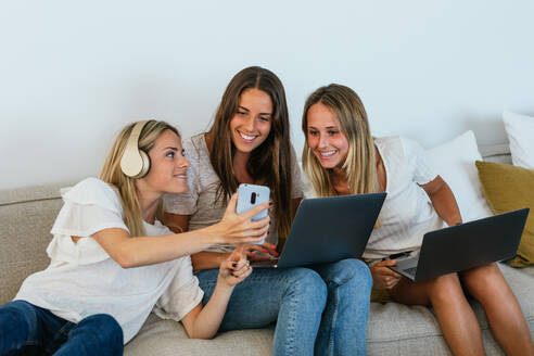 Frau mit Kopfhörern zeigt fröhlichen Freundinnen mit Laptops ein Video auf dem Handy, während sie auf der Couch im Wohnzimmer sitzen - ADSF34284