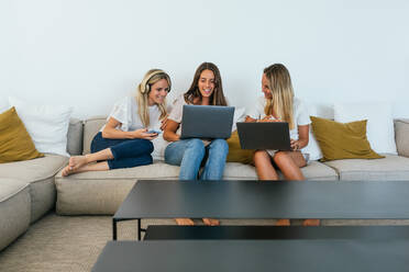 Woman showing a video on laptop to cheerful friends sitting around her on the living room sofa - ADSF34285