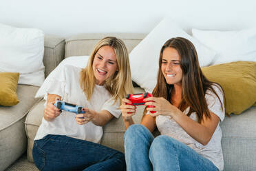 Bild von positiven Freundinnen mit Gamecontrollern, die ein Videospiel spielen, während sie auf dem Boden sitzen und sich auf das Sofa im Wohnzimmer lehnen - ADSF34291
