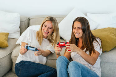 Crop of positive female friends with game controllers playing videogame while sitting on the floor leaning on the sofa in living room at home - ADSF34291