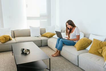 Seitenansicht einer konzentrierten Frau in Freizeitkleidung, die auf einem Netbook tippt und telefoniert, während sie auf einem bequemen Sofa mit Kissen im hellen Wohnzimmer zu Hause sitzt - ADSF34295