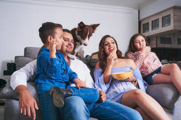 Positive Familie mit einer Schüssel Essen auf der Couch sitzend mit einem Border Collie Hund, während sie im hellen Wohnzimmer fernsehen - ADSF34299