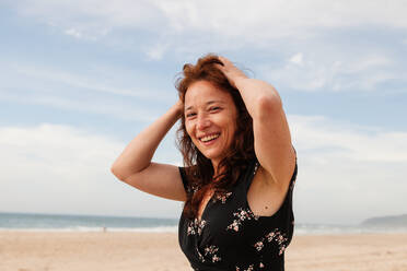 Positive female in black sundress touching hair while standing against cloudy sky in nature on summer day - ADSF34309