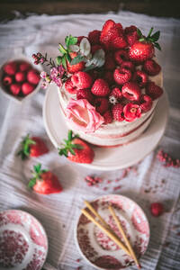 From above delicious red velvet cake topped with fresh strawberries and decorated with flowers served on table with white tablecloth in kitchen - ADSF34337