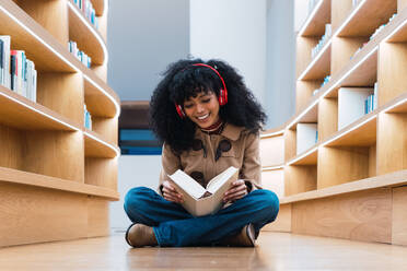 Glückliche afroamerikanische Studentin mit Kopfhörern, die ein Buch liest, während sie in der Bibliothek auf dem Boden sitzt und genießt - ADSF34345