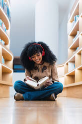 Glückliche afroamerikanische Studentin mit Kopfhörern, die ein Buch liest, während sie in der Bibliothek auf dem Boden sitzt und genießt - ADSF34346