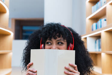 Schwarze Frau mit lockigem Haar, die mit einem aufgeschlagenen Buch gegen ein verschwommenes Bücherregal in einer Bibliothek schaut, während sie Musik hört - ADSF34347