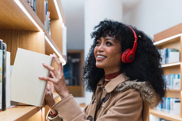 Seitenansicht einer positiven afroamerikanischen Frau, die Musik hört und ein Buch aus einem Bücherregal in der Bibliothek nimmt - ADSF34348