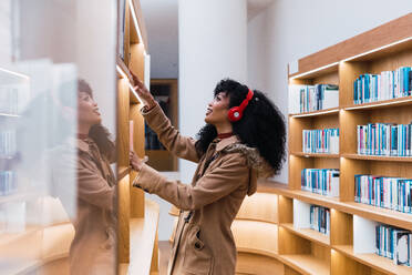 Seitenansicht einer positiven afroamerikanischen Frau, die Musik hört und ein Buch aus einem Bücherregal in der Bibliothek nimmt - ADSF34349