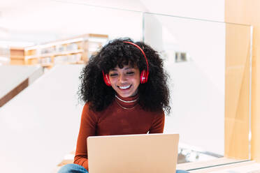 Schwarze, lockige, fröhliche Frau mit Kopfhörern, die mit einem Laptop in einer modernen Bibliothek sitzt - ADSF34351