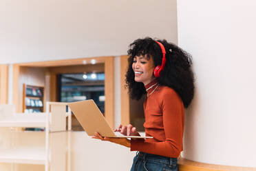Schwarze, lockige, fröhliche Frau mit Kopfhörern, die mit einem Laptop in einer modernen Bibliothek steht und sich an die Wand lehnt - ADSF34352
