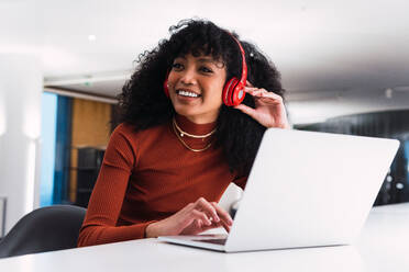 Inhalt Afroamerikanische Frau mit Kopfhörern, die telefoniert, sitzt an einem Tisch mit Laptop in einem modernen Arbeitsraum - ADSF34355