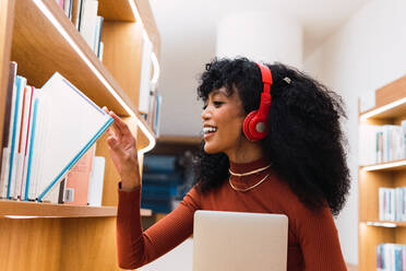 Seitenansicht einer positiven afroamerikanischen Frau, die Musik hört und ein Buch aus einem Bücherregal in der Bibliothek nimmt - ADSF34356