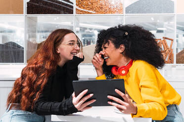 Seitenansicht von fröhlichen jungen multiethnischen weiblichen besten Freunde in lässiger Kleidung lachen fröhlich beim Sitzen in modernen Café mit Tablet-Computer in den Händen - ADSF34363