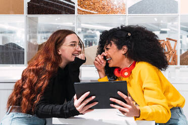 Side view of cheerful young multiracial female best friends in casual clothes laughing happily while sitting in modern cafe with tablet computer in hands - ADSF34363