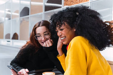 Seitenansicht von fröhlichen jungen multiethnischen besten Freundinnen in legerer Kleidung, die glücklich lachen, während sie in einem modernen Café mit Smartphones in den Händen sitzen - ADSF34367
