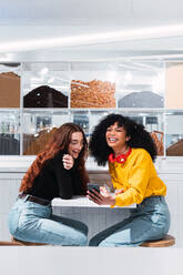 Seitenansicht von fröhlichen jungen multiethnischen besten Freundinnen in legerer Kleidung, die glücklich lachen, während sie in einem modernen Café mit Smartphones in den Händen sitzen - ADSF34368