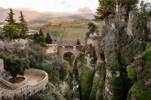 Breathtaking landscape of old stone arched bridge over river surrounded by massive rocky cliffs against cloudy sunset sky in Ronda - ADSF34383