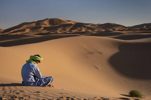 Ein Berber in traditioneller Kleidung sitzt inmitten der Sanddünen von Erg Chebbi, Sahara-Wüste, Marokko, Nordafrika, Afrika - RHPLF21973