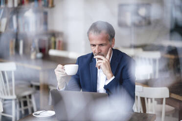 Geschäftsmann mit Hand am Kinn und Kaffeetasse am Laptop in einem Café - GUSF07204