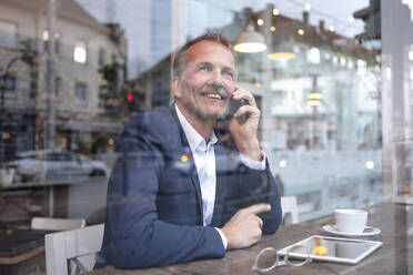 Lächelnder Geschäftsmann, der mit seinem Handy telefoniert und durch das Fenster eines Cafés schaut - GUSF07205