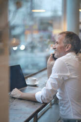 Businessman looking through window and talking on mobile phone at cafe - GUSF07209