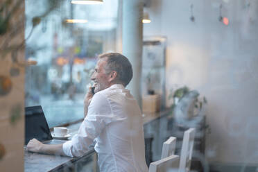 Smiling businessman sitting by glass window and talking on mobile phone - GUSF07212