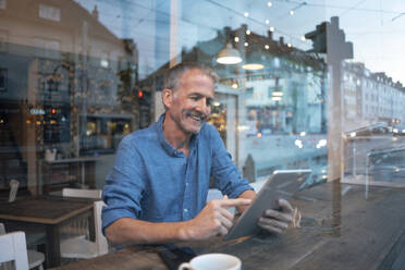 Lächelnder Geschäftsmann sitzt an einem Tisch vor einem Fenster und benutzt einen Tablet-PC in einem Café - GUSF07217