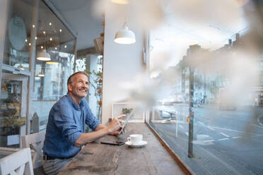 Glücklicher Geschäftsmann mit Tablet-PC, der am Tisch vor einem Glasfenster in einem Café sitzt - GUSF07218
