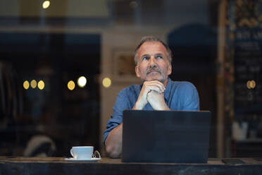 Thoughtful businessman with laptop and coffee cup sitting at table in cafe - GUSF07231