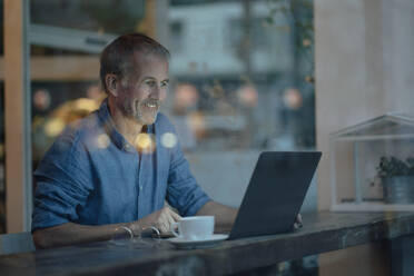 Lächelnder Geschäftsmann mit Laptop und Kaffeetasse, der an einem Tisch am Glasfenster in einem Café sitzt - GUSF07232