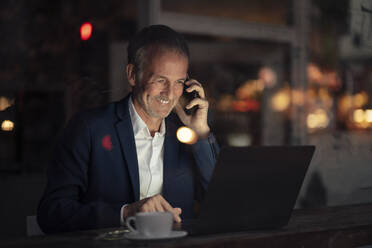 Happy businessman talking on mobile phone and using laptop at night - GUSF07242